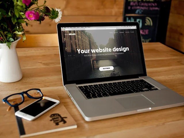Laptop on a wooden desk displaying a Your website design homepage, beside flowers, glasses and a smartphone on a notebook