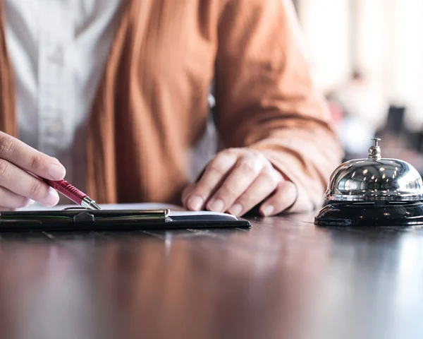 Guest in a tan cardigan writing at a hotel reception desk with a chrome call bell, linked to taking reservations online