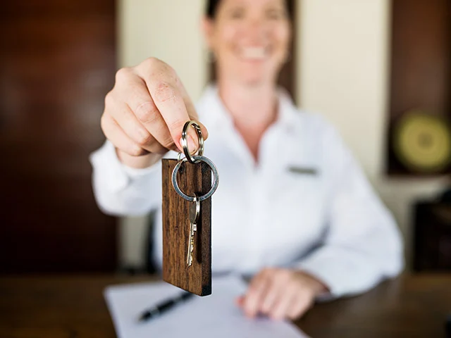 Smiling receptionist in a white shirt holding out a room key on a wooden tag, showing a booking completed at check-in