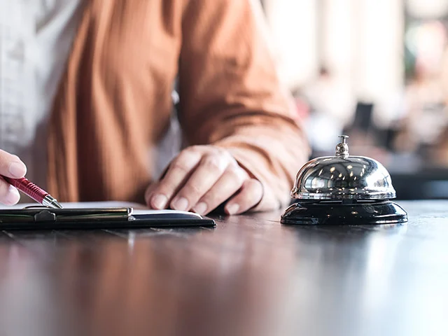Pen filling in a form at a wooden front desk next to a silver service bell, representing more direct bookings for a hotel