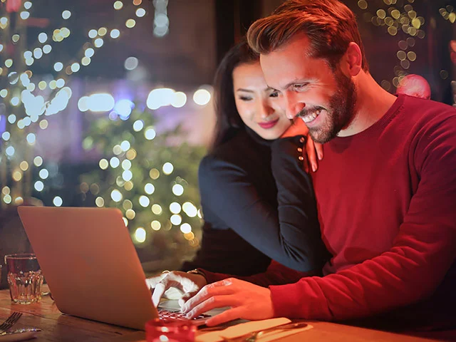 Smiling couple using a laptop at a cafe table with twinkling lights, showing an easy online hotel booking experience