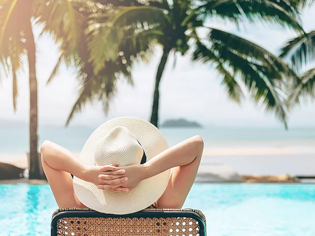 Woman in a straw sun hat relaxing by an infinity pool facing palm trees and the sea, the kind of stay a direct offer sells
