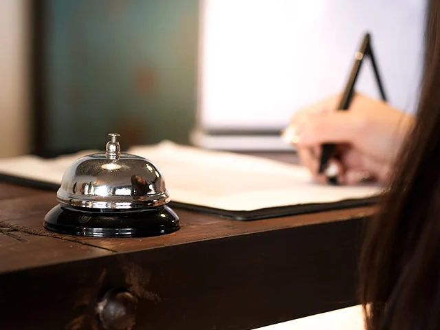 Hotel receptionist writing in a ledger behind a silver service bell on a wooden front desk, taking a direct booking