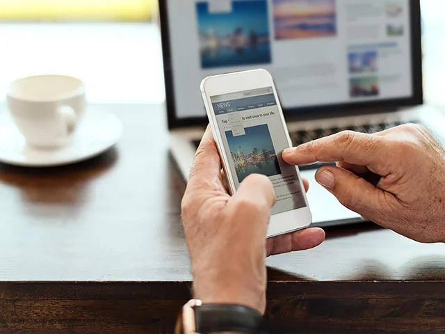 Man reading a travel news site on a smartphone with a laptop and coffee cup behind, showing mobile-friendly design