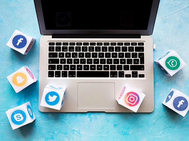 Laptop on a blue surface surrounded by cubes with Facebook, Twitter, Instagram, Skype, WhatsApp and Snapchat logos