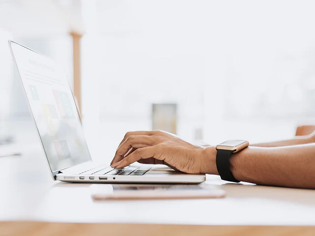 Person wearing a smartwatch typing on a laptop at a bright white desk beside a phone, working on hotel online marketing