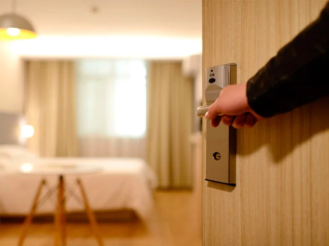 Guest opening a hotel room door fitted with a key card lock, a softly lit bed and curtained window inside