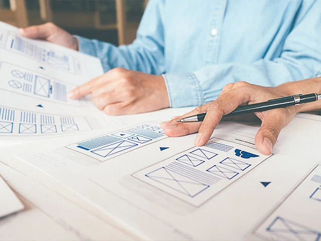 Hand holding a pen over paper website wireframes drawn in simple blue lines, planning a clean, minimal layout