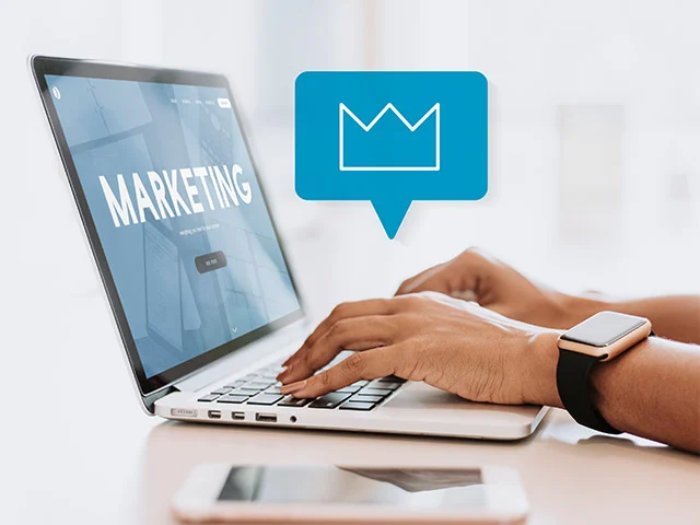 Hands typing on a laptop showing the word Marketing beside a blue crown icon, using content to win direct bookings