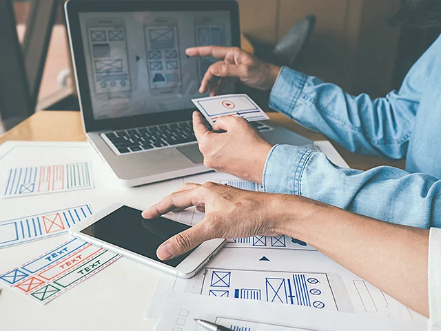 Designer tapping a tablet and holding a mobile wireframe card in front of a laptop, planning a responsive hotel site