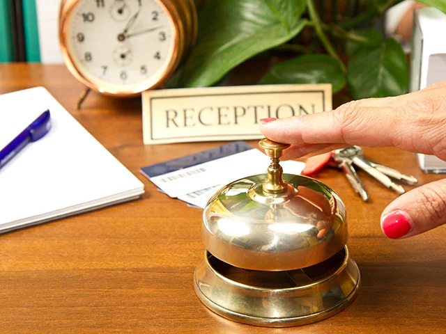 Finger pressing a brass bell on a desk with a Reception sign, key cards, room keys and an alarm clock
