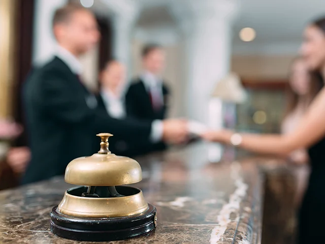 Brass service bell on a marble hotel reception desk while a guest shakes hands with staff at check-in