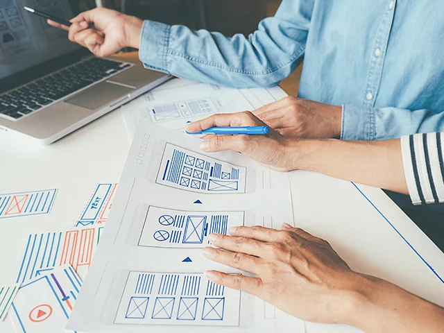 Two designers reviewing paper wireframes with a blue pen, one pointing at a laptop, sketching a hotel website layout