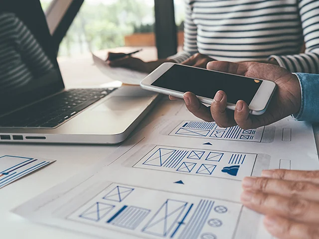 Hand holding a smartphone over paper wireframe sketches beside a laptop, planning hotel website improvements