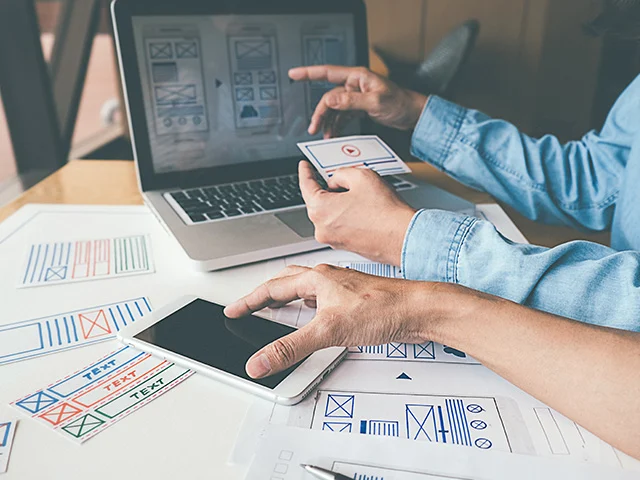 Designer in a denim shirt holding wireframe cards by a laptop and smartphone, planning a hotel website