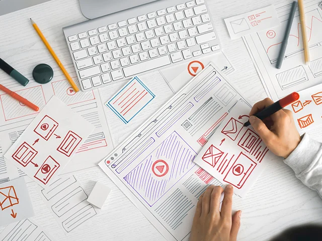 Designer drawing red website wireframes with a marker on paper sketches beside a keyboard and pencils