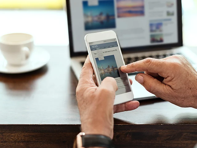 Hands scrolling a travel news page on a smartphone in front of a laptop and coffee cup