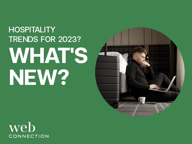 Man in black sitting on a hotel room floor with a laptop and coffee cup beside a suitcase, next to text asking What's New