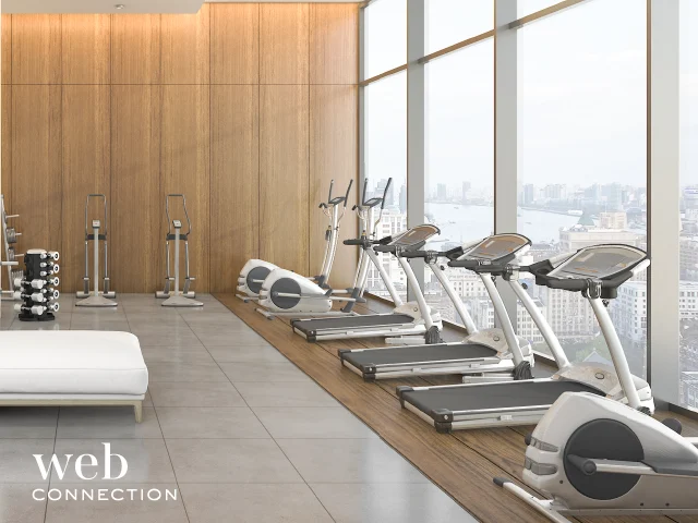 Bright hotel gym with treadmills and elliptical trainers lined along floor-to-ceiling windows overlooking a city river
